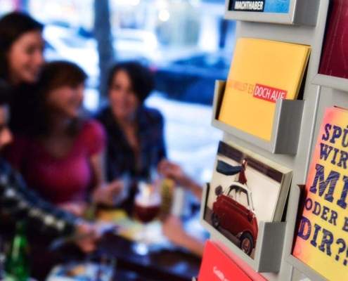 Bunter Postkartenständer mit humorvollen Motiven und Sprüchen in einem Café, mit einer Gruppe von Freunden im Hintergrund, die entspannt zusammen sitzen.