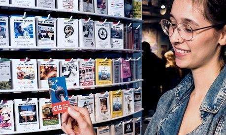 minicard-werbung-berlin-tourismus-rabattkarten Eine Frau nimmt eine Minicard mit Rabattangebot aus einem Displayständer – Beispiel für touristische Vorteils- und Werbekarten in Berlin.