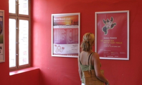 plakatwerbung-kulturveranstaltung-gastronomie-innenraum Frau betrachtet bunte Plakate an einer roten Wand, die für kulturelle Veranstaltungen und Gastronomieangebote werben – Beispiel für wirkungsvolle Innenraumwerbung und Plakatpromotion.